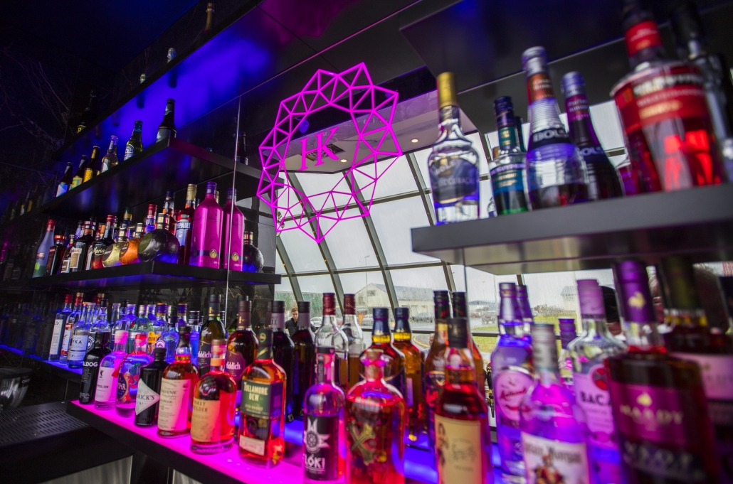 A bar shelf is stocked with various bottles of liquor and spirits displayed in rows. Bright LED lighting, including pink illumination and a geometric neon sign with "IK" on it, evokes the stylish ambiance of a luxury Iceland break.