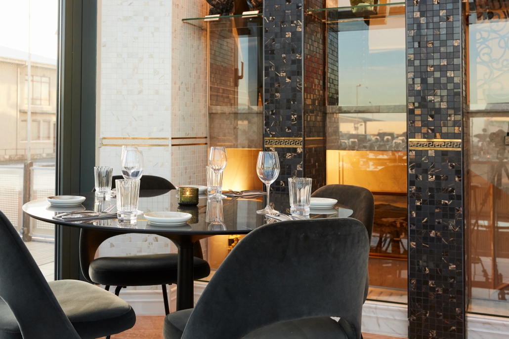 A modern dining table set for four with glassware and plates sits in a chic restaurant. The backdrop features tall, decorative black and golden columns from Juliettes Trade with a reflective surface, and large windows reveal an urban exterior with soft sunlight.