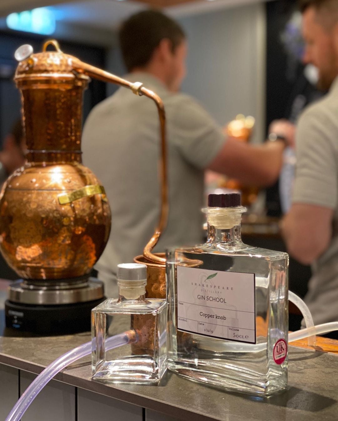 A copper still apparatus is displayed on a counter next to two glass bottles labeled "GIN SCHOOL" and "Copper Knob." In the background, two individuals, perhaps enjoying a staycation activity, are engaged in conversation, slightly out of focus. The scene appears to be inside a distillery or bar.