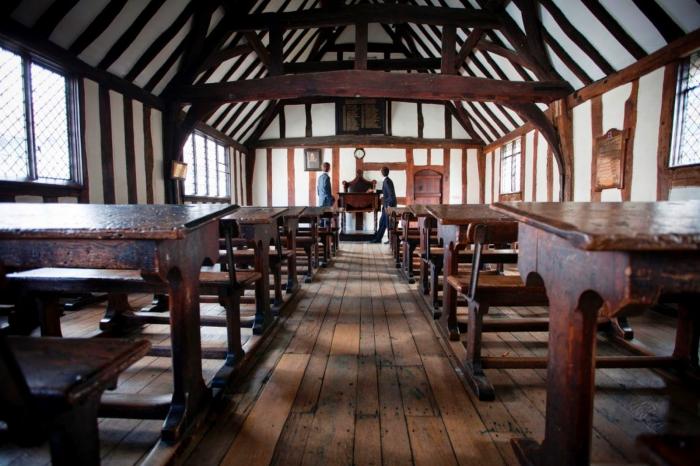 A historic classroom with wooden desks and benches aligns symmetrically on either side of a central aisle. The room features wooden beams, white walls, leaded glass windows, and a high ceiling. It’s an ideal spot for a nostalgic staycation. Two people are standing at a podium near the far end of the room.