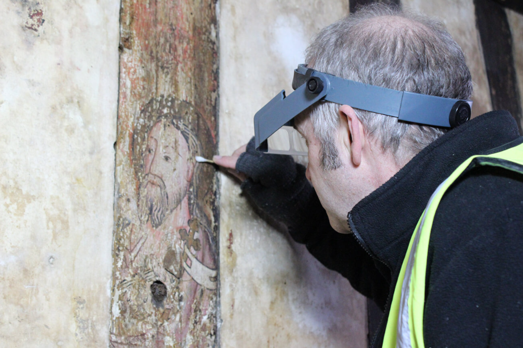 A person wearing a visor and high-visibility vest is diligently working on restoring a historic wall painting of a bearded figure in a crown. With a small tool in hand, they carefully clean or repair the artwork, highlighting the delicate nature of the process—a staycation at its finest for art enthusiasts.