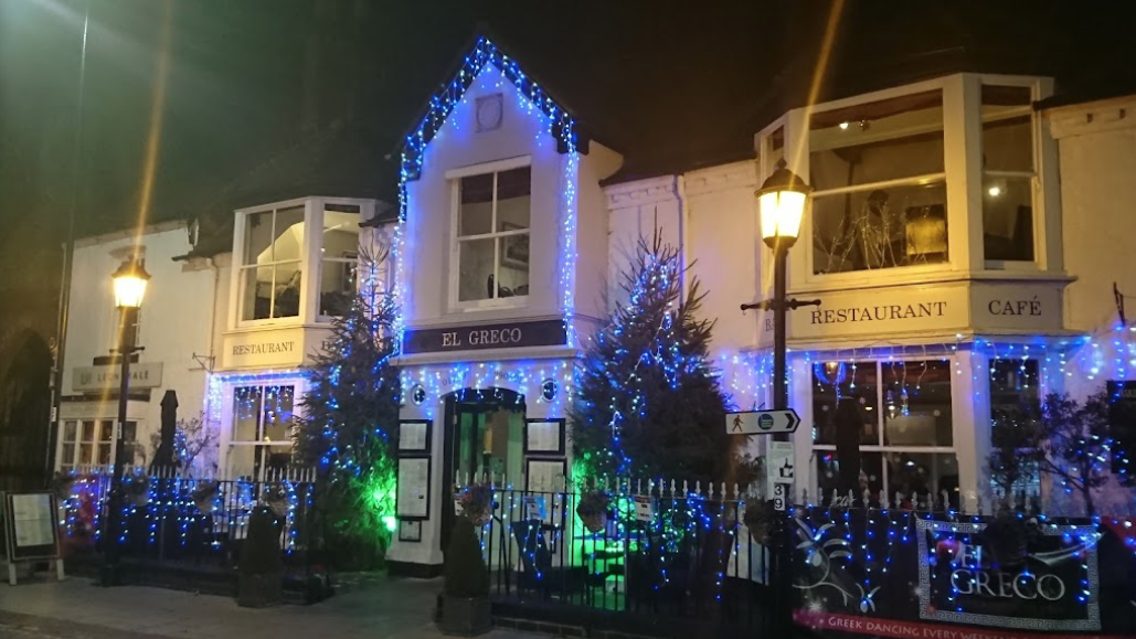 A cozy restaurant named "El Greco" is adorned with festive blue and green lights, along with several Christmas trees. The building is white with dark trimmings and has large windows. It is night, and the area is illuminated by street lamps, making it an ideal staycation spot for holiday cheer.