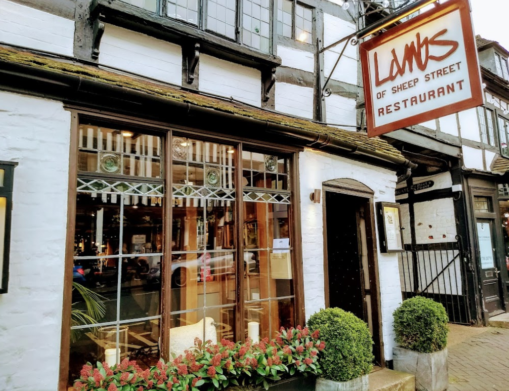 A historic building with a wooden facade, housing "Lambs of Sheep Street" restaurant, perfect for a charming staycation. The entrance is framed by large windows, potted plants, and a brown hanging sign. Inside, the interior is warmly lit and inviting. Streetscape reflections are visible in the glass.