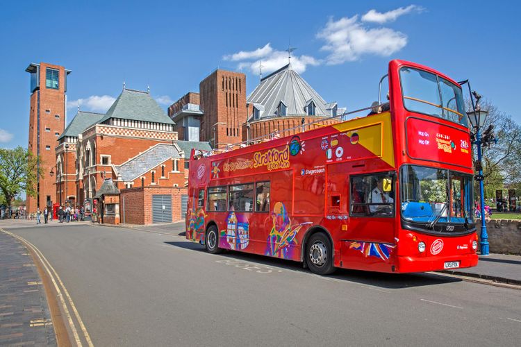 A red double-decker tour bus is parked on a street with a historical building and a clock tower in the background. The bus features colorful graphic advertisements and the word "Stratford" on its side. Tourists enjoying their staycation are seen walking nearby on a clear, sunny day.