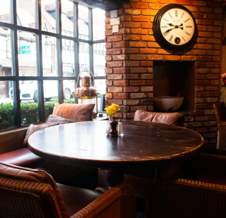 A cozy café interior with a round wooden table surrounded by wicker chairs invites the perfect staycation. A single yellow rose in a vase sits on the table, while a brick wall features a large clock. Tall windows provide a view of parked cars and buildings outside, and warm lighting enhances the ambiance.