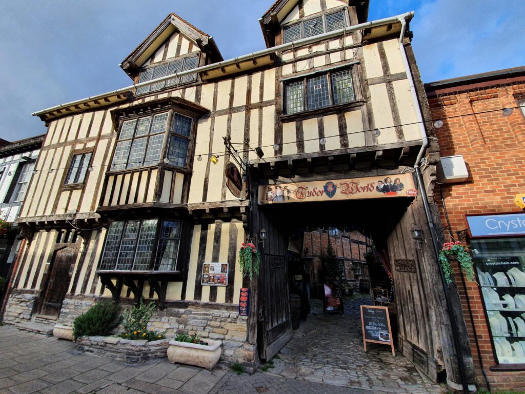 A historical building with timber-framed architecture and overhanging upper floors. A sign reads "The Crooked House" above an arched passageway leading to a courtyard filled with plants. Perfect for a staycation, the building is flanked by a brick structure on the right.