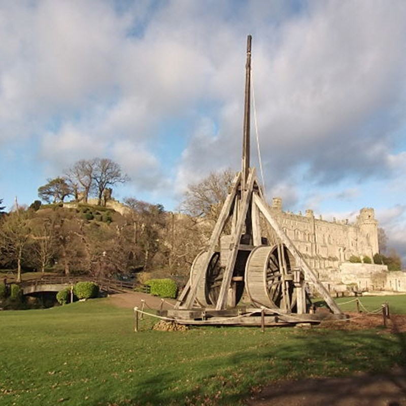 A large wooden trebuchet is set up on a grassy lawn in front of a historic stone castle, creating the perfect backdrop for your staycation. Trees and a hill are visible in the background under a partly cloudy sky. A wooden fence surrounds the trebuchet, and a stone pathway leads towards the castle.