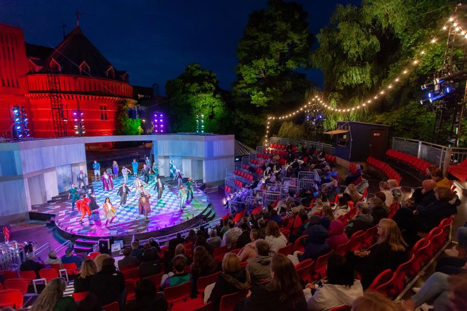 A vibrant outdoor theater performance takes place at night, offering the perfect staycation activity. A well-lit stage features actors in colorful costumes as the audience sits in red seats under strings of lights. Trees and an illuminated building are visible in the background.