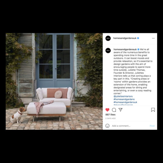 A cozy outdoor seating area features a plush pink-and-white daybed with cushions and a throw blanket. Lush greenery climbs the adjacent wall, framing a rustic blue door. A small brown-and-white dog stands nearby, looking towards the camera. An Instagram post overlay is visible.