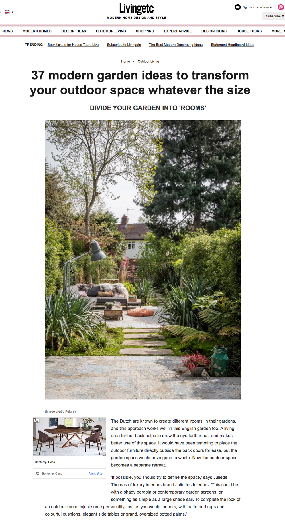 A garden with lush greenery, a stone pathway, and a cozy seating area surrounded by plants. There is a mix of tall trees, shrubs, and flowering plants, creating a serene outdoor space. A brick house with a patio is visible in the background.