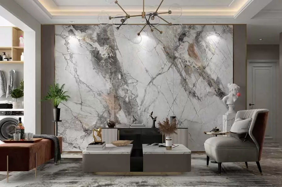 A modern living room features a large white marble accent wall, grey and white furnishings, and a contemporary chandelier. A statue accents a corner near a grey chair, while a coffee table with vases and plants, and a leather bench complete the stylish decor.