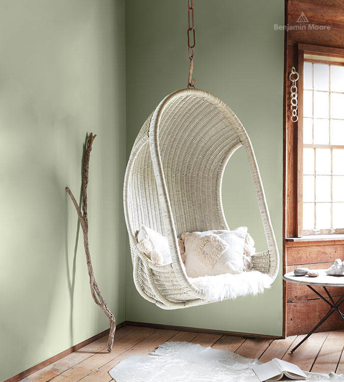 A cozy, minimalist room with sage-green walls, reflecting colour trends 2022, features a rattan hanging chair with fluffy white cushions and throws. Natural wooden floors and trim, along with a window letting in light, add warmth. A decorative branch leans against the wall, enhancing the serene ambiance.