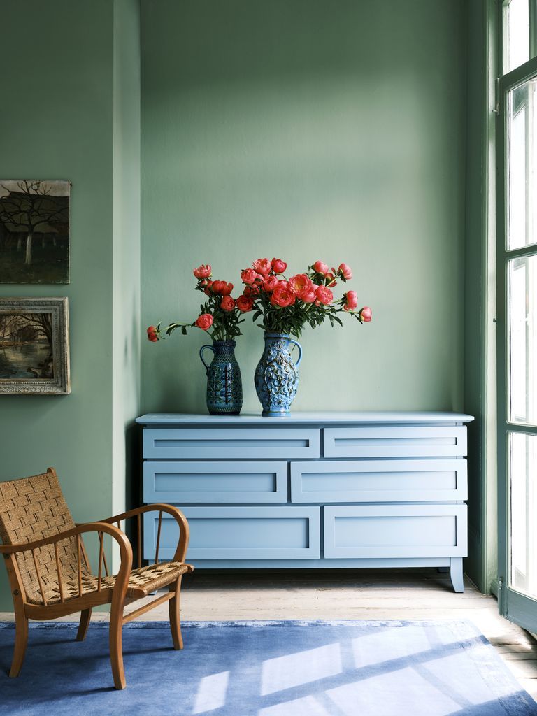 A light blue wooden dresser sits against a light green wall, topped with two blue vases filled with vibrant pink flowers. To the left, there's a wicker chair with a woven seat, and part of a framed painting is visible. A large window on the right lets in natural light, reflecting colour trends 2022.