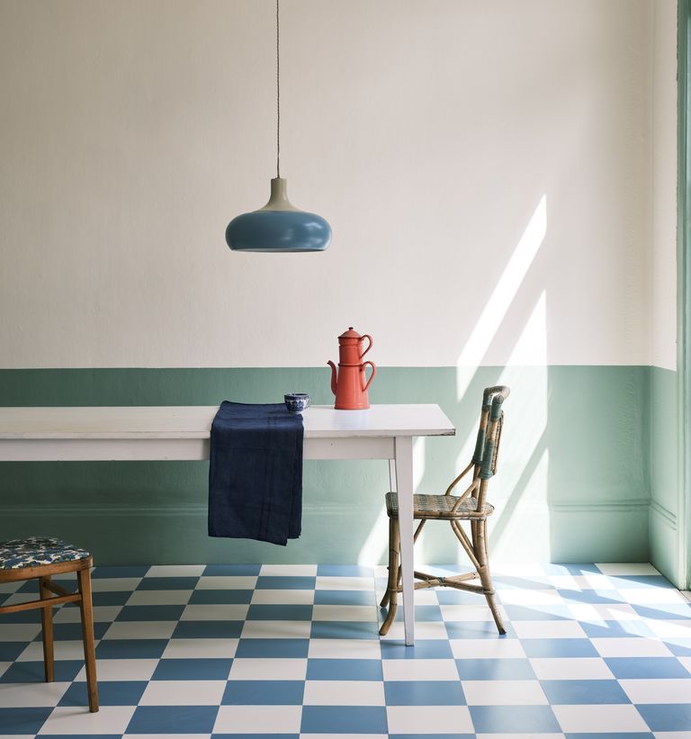 A minimalist dining room features a rectangular white table with a blue cloth and an orange coffee pot. Two mismatched chairs sit on either side, and a blue pendant light hangs above. Reflecting the colour trends of 2022, the room has green lower walls and checkered blue-and-white flooring.