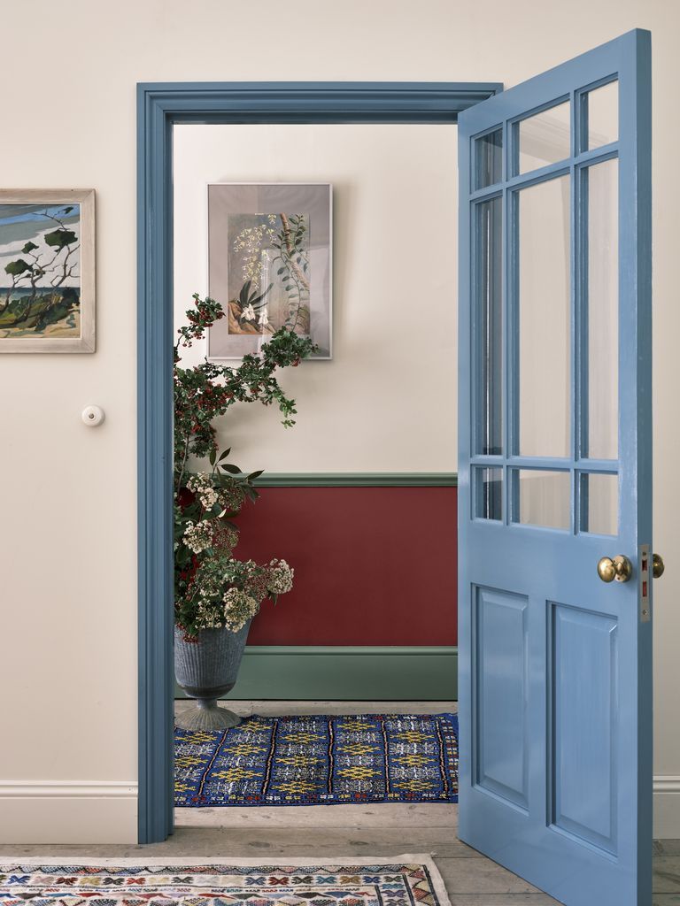 A light blue door is partially open, revealing a hallway with an intricate patterned rug, a red wainscoting with green trim, and potted plants. Two framed artworks hang on the beige walls, showcasing color trends 2022 and adding an artistic touch to the cozy entryway.