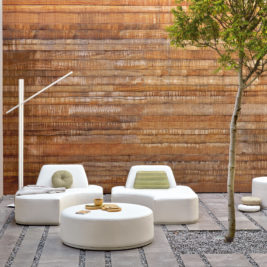 Modern outdoor patio with white furniture, including round and rectangular seating and tables, arranged on a stone-paved floor with pebbled lines. Reflecting garden trends 2022, a single green tree provides shade, while a tall, minimalist white floor lamp stands nearby. A rust-colored wall serves as the backdrop.