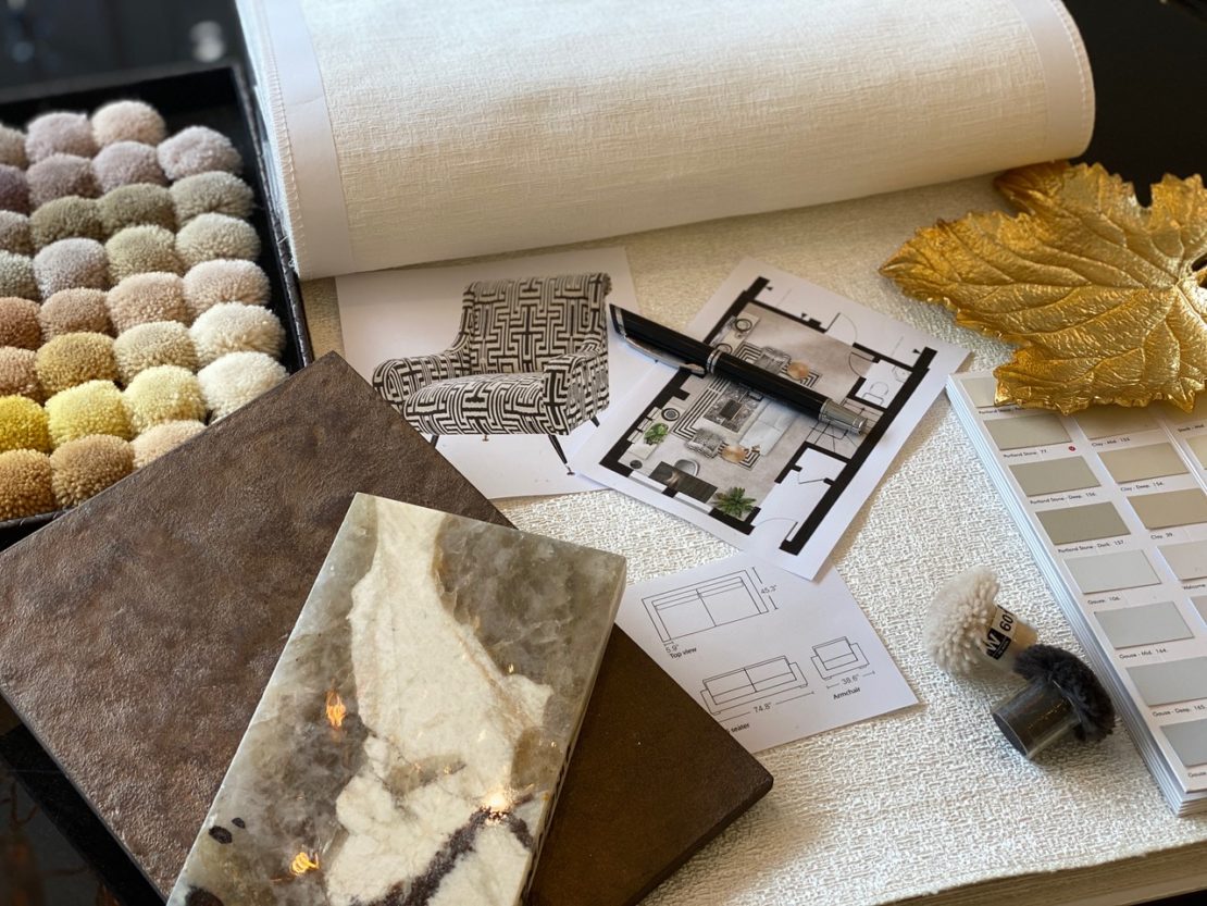 An interior design workspace featuring fabric and carpet samples, a white textured roll, a pen, architectural plans, a golden leaf decoration, and material swatches. The scene invites you to become your own interior designer by showcasing various elements of the design and planning process.