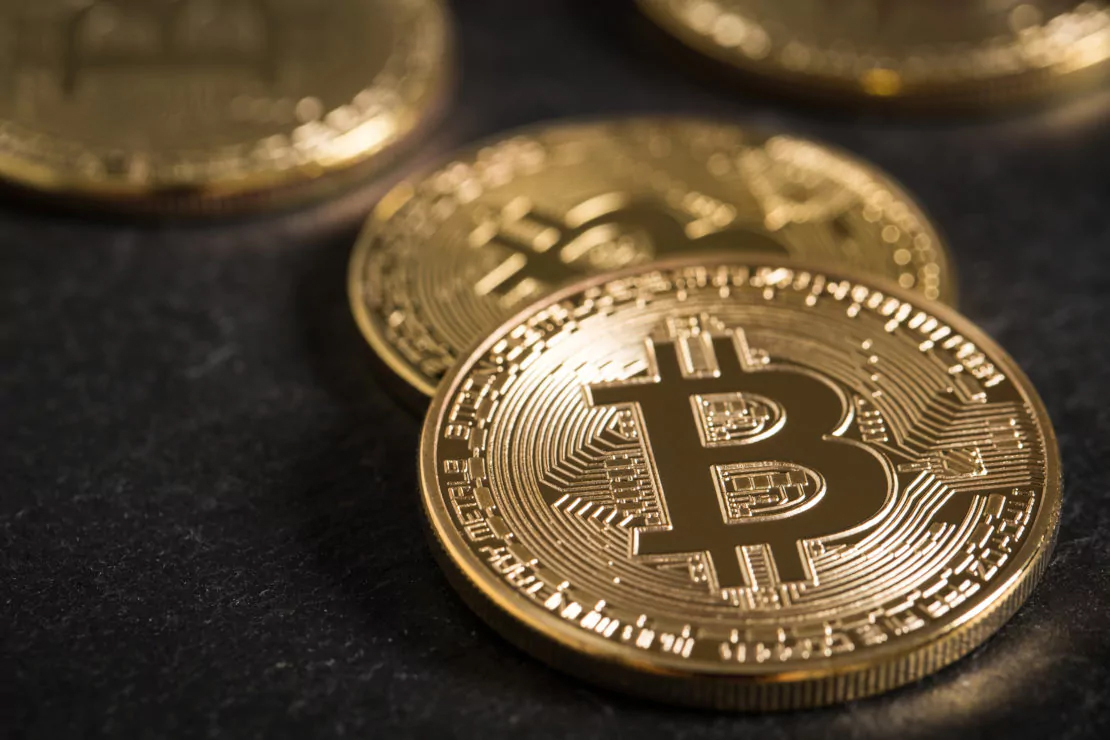 A close-up of several gold-colored Bitcoin coins placed on a dark surface. The foreground coin prominently displays the Bitcoin symbol (₿), with intricate circuit-like patterns surrounding it. Other coins are partially visible in the background, reflecting a sharp increase in crypto transactions by luxury spenders.