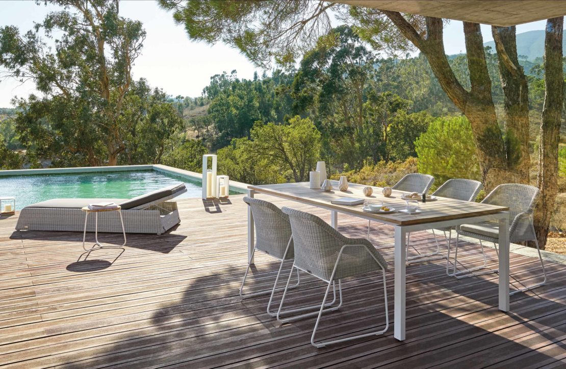 A serene outdoor deck featuring a dining table set with four chairs and minimalistic decor, perfect for al-fresco dining ideas for your garden. Beside the deck is a large swimming pool bordered by lush, green landscape with tall trees. A single lounge chair and side table are placed beside the pool for relaxation.