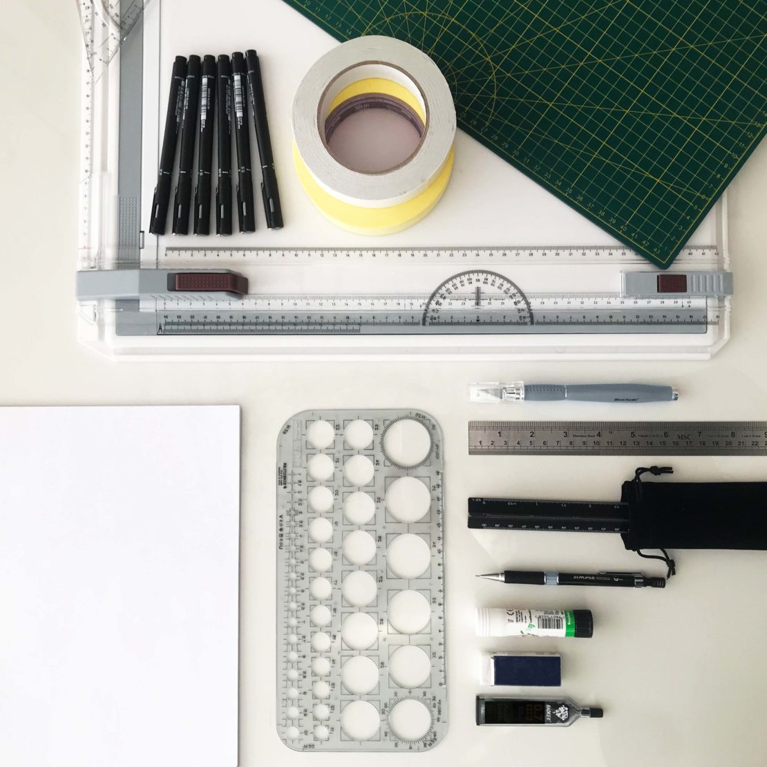 Become your own interior designer with this arrangement of drafting tools including a drawing board, a green cutting mat, tape, pens, a compass, a ruler, a circle template, an eraser, and correction fluid on a white surface.