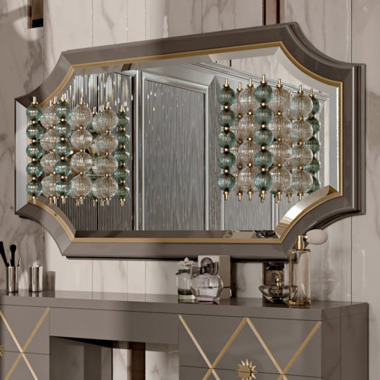 A chic, modern vanity setup featuring a large, decorative mirror with a gold-trimmed, geometric frame that echoes art deco interior design. The mirror reflects an elaborate light fixture made of multiple glass orbs. The vanity table below has elegant patterned drawers and assorted grooming items.