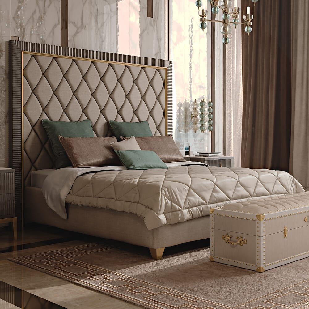 A luxurious bedroom features a tufted, upholstered headboard and a neatly made bed with beige bedding and decorative pillows in muted green and brown. The room, showcasing art deco interior design, is accented with marbled walls, elegant lighting fixtures, heavy drapes, and a vintage-style storage chest at the foot of the bed.