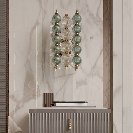 A modern interior scene showcasing a wall-mounted light fixture with several spherical, textured glass bulbs in green and clear colors. The light, reflecting art deco interior design elements, is set against a marble-patterned wall above a gray cabinet with fluted texture and brass accents.