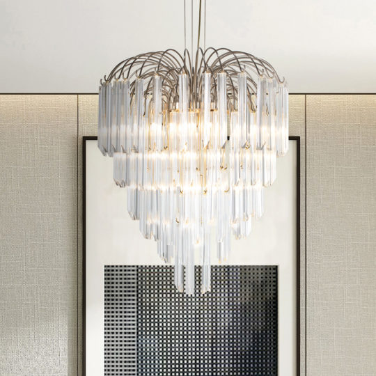 A modern chandelier with multiple cascading glass or crystal rods, emitting a warm glow, hangs from the ceiling. The fixture is positioned in front of a rectangular framed print with a geometric design on a textured wall, echoing the elegance of art deco interior design.