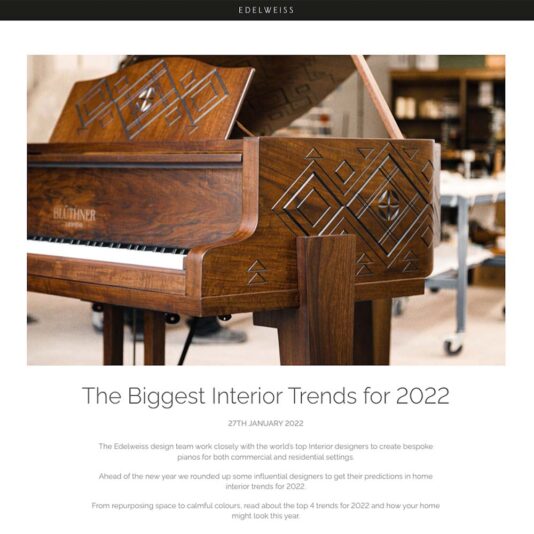 A beautifully crafted wooden piano with intricate geometric carvings on its side is the focal point of the image. Above the piano is the title "The Biggest Interior Trends for 2022," followed by a short description about design team work and trends.