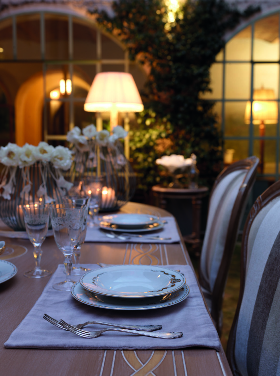 A beautifully set dining table outdoors during the evening. The table features elegant dinnerware, glassware, and cutlery on a light gray placemat. Soft, decorative lighting and white flower arrangements add a warm, inviting ambiance to the scene—perfect for al-fresco entertaining tips.