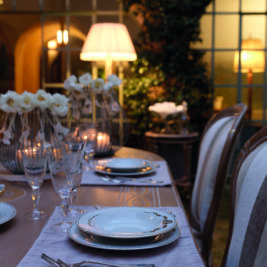 A beautifully set dining table outdoors during the evening. The table features elegant dinnerware, glassware, and cutlery on a light gray placemat. Soft, decorative lighting and white flower arrangements add a warm, inviting ambiance to the scene—perfect for al-fresco entertaining tips.