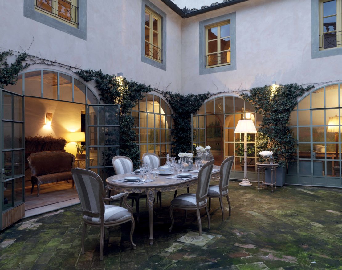 A cozy courtyard for al-fresco entertaining tips features a round dining table set with plates and glassware surrounded by elegant chairs. The space is enclosed by arched windows and doors, each adorned with climbing ivy. Warm lighting from lamps and wall fixtures creates an inviting ambiance.