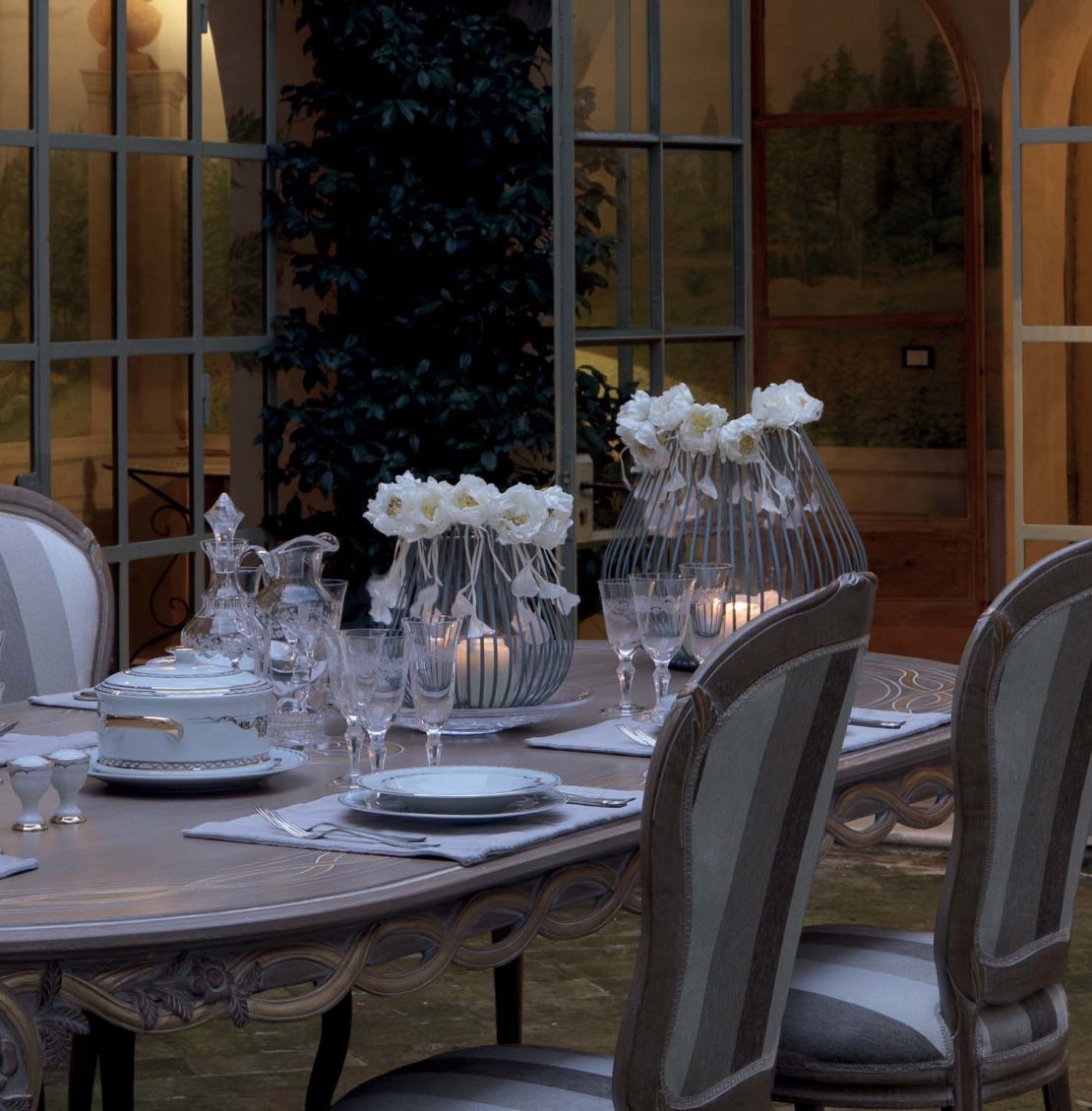 A stylish evening table setting with elegant, muted-tone chairs around a wooden table adorned with white flowers in vases, crystal glassware, and fine china. The backdrop features large windows, a painted mural, and soft lighting, offering al-fresco entertaining tips for an inviting ambiance.