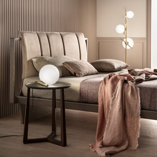 A contemporary bedroom showcases a wooden bed with beige upholstery, accented by beige and pink bedding. Beside the bed, a small round side table holds a spherical white lamp. In the corner, the Modern Murano Glass Floor Lamp, featuring three spherical lights, enhances the room's warm, neutral tones.