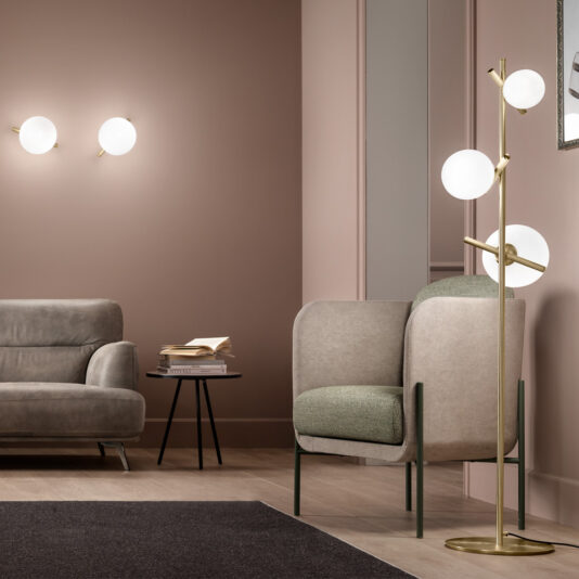 A modern living room with muted pink walls featuring two wall-mounted round lights, a gray sofa, an armchair with green and beige upholstery, and a stylish Modern Murano Glass Floor Lamp showcasing three globe lights. A small round side table with stacked books adds a cozy touch.