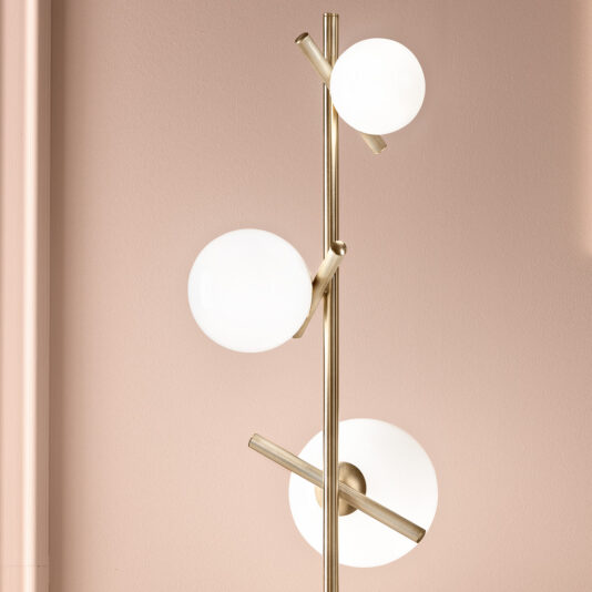 The Modern Murano Glass Floor Lamp boasts a gold metallic stand with three white spherical bulbs arranged at varying heights. Its sleek and contemporary design is elegantly showcased against a light pink wall.