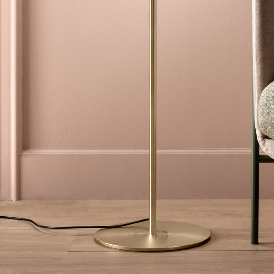 A close-up of the base of a Modern Murano Glass Floor Lamp, featuring a sleek, metallic gold stand. The lamp is placed next to a padded chair adorned with a textured cushion, while a black power cable lies on the light-colored wooden floor. The background wall is painted in soft pink tones.