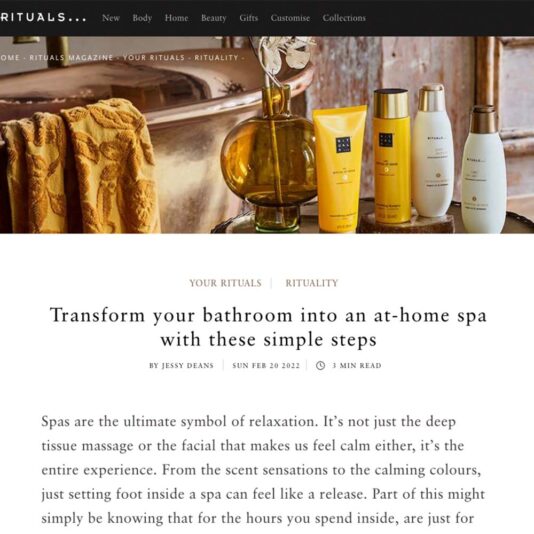 A selection of Rituals body care products, including lotions and oils, is displayed on a wooden shelf with a yellow towel hanging nearby. The background features a wooden bathroom setting, contributing to the serene and spa-like atmosphere Rituals is known for.