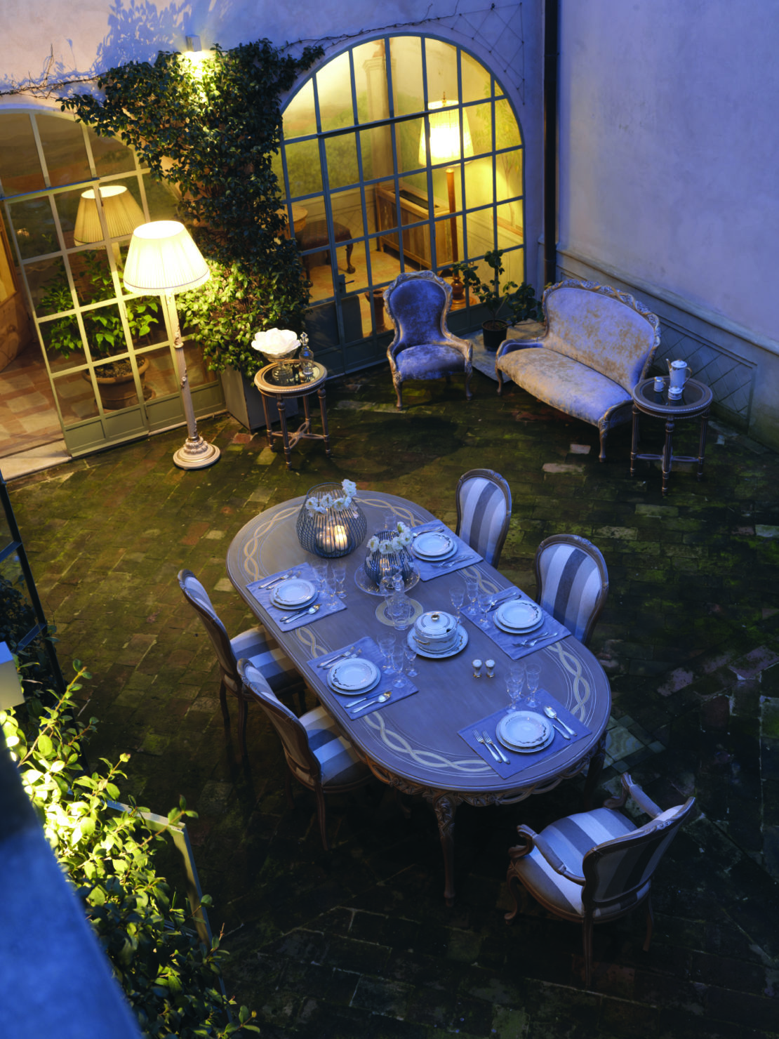 An outdoor dining area with an elegant oval table set for a meal, featuring ornate chairs and tableware. The surrounding courtyard, ideal for al-fresco entertaining tips, has lush greenery, vintage armchairs, a sofa, a side table, and ambient lighting from a floor lamp, creating a cozy and inviting atmosphere.