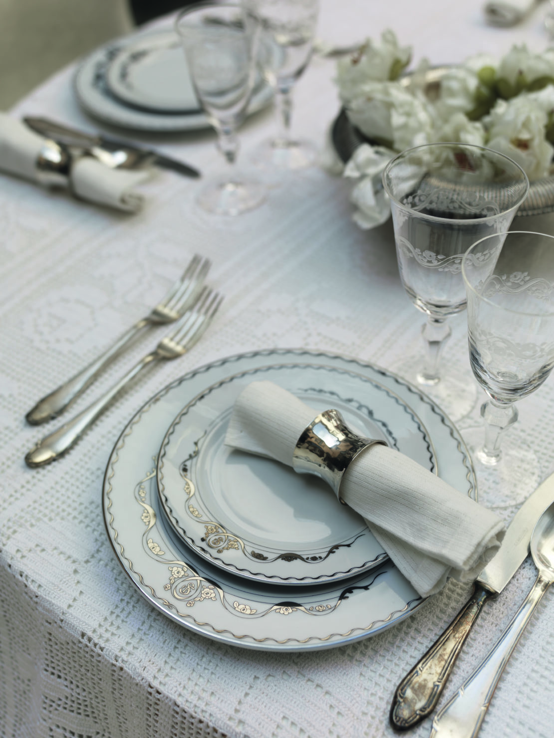 Elegant table setting with fine china plates stacked atop a lacy white tablecloth. Silverware and crystal glasses surround the plates, with a white napkin in a silver holder on top. A floral centerpiece with white flowers adds a touch of sophistication—perfect for al-fresco entertaining tips.