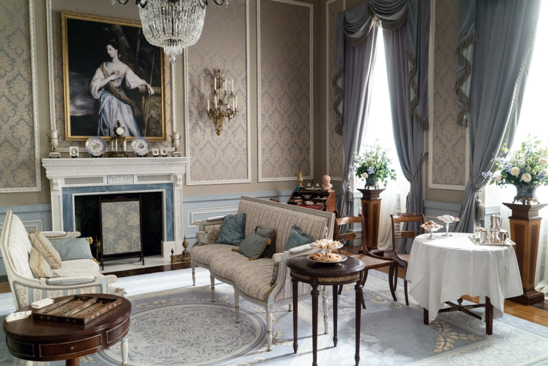 An elegant Victorian-era room with ornate wallpaper and high ceilings features antique furniture, including sofas, armchairs, and small tables. A large portrait hangs above a grand fireplace. The room is adorned with drapes, flowers, and a chandelier—perfect for those seeking Bridgerton-inspired home ideas.