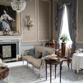 An elegant Victorian-era room with ornate wallpaper and high ceilings features antique furniture, including sofas, armchairs, and small tables. A large portrait hangs above a grand fireplace. The room is adorned with drapes, flowers, and a chandelier—perfect for those seeking Bridgerton-inspired home ideas.