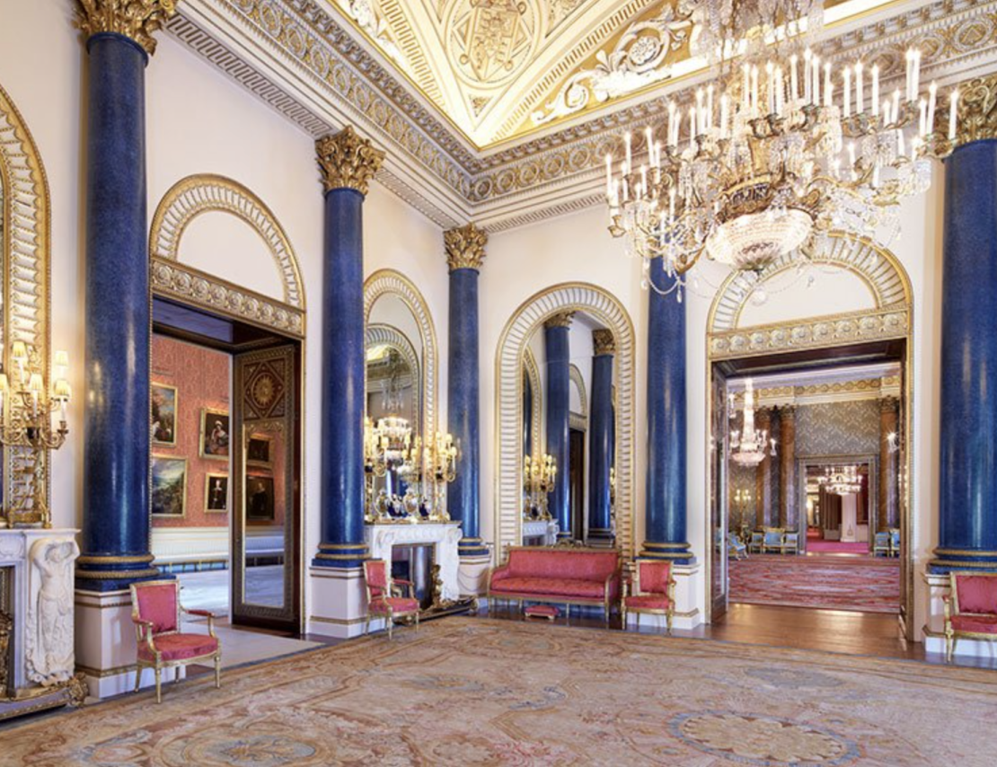 An opulent ballroom with tall blue columns, gilded trim, grand mirrors, and luxurious décor reminiscent of the grandest rooms of Queen Elizabeth. Elegant chandeliers hang from the intricately designed ceiling. Ornate red and gold furniture is spaced around the room, with paintings adorning the walls in the background.