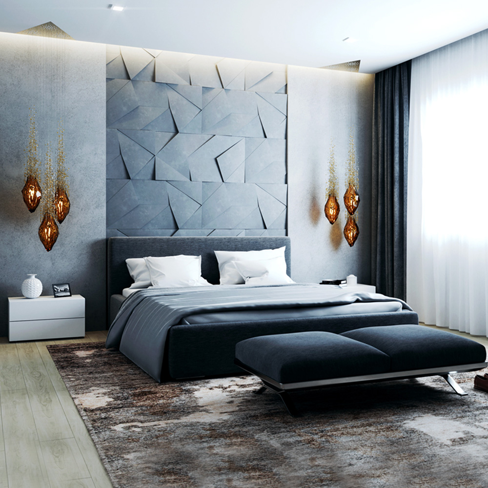 A modern bedroom features a large bed with a gray headboard against a textured geometric wall. The bed has white and gray linens. Sculptural lighting that instantly transforms your home hangs on each side in the form of amber glass pendant lights. A matching gray bench sits at the foot of the bed, alongside white cabinets and thick drapes.