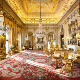 A grand, opulent room reminiscent of the grandest rooms of Queen Elizabeth, with intricate gold-plated designs on the ceiling and walls. The space features large chandeliers, ornate furniture, and a spacious red carpet with detailed patterns. Tall windows allow natural light to enhance the luxurious ambiance.