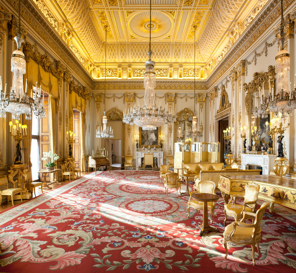 A grand, opulent room reminiscent of the grandest rooms of Queen Elizabeth, with intricate gold-plated designs on the ceiling and walls. The space features large chandeliers, ornate furniture, and a spacious red carpet with detailed patterns. Tall windows allow natural light to enhance the luxurious ambiance.