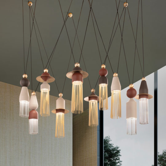 A collection of Contemporary Brown LED Pendant Light Clusters hangs from the ceiling, featuring unique, artistic designs. The LED lights come in various geometric shapes in muted colors, adorned with fringes and suspended at different heights to create a captivating display. Through a window in the background, some foliage can be seen outside.
