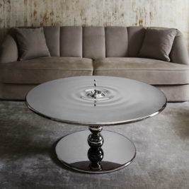 A circular coffee table with a reflective, metallic surface that resembles rippling water stands in front of a gray velvet sofa. The background includes a textured, neutral wall, and the setting exudes a modern, minimalistic aesthetic. Make a statement with these luxury table designs.