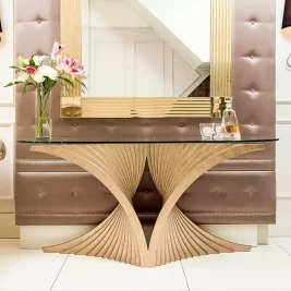 A luxurious hallway features a gold sculptural console table with a glass top, adorned with a vase of fresh flowers and a glass decanter set. Behind the table, a large, ornate gold-framed mirror hangs on a tufted leather wall panel.