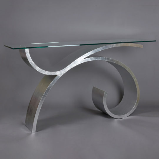 A modern console table featuring a sleek, curved metallic base and a glass top. The base consists of a large, artistic swirl design, giving the table a contemporary and artistic appearance against a plain gray background—an ideal choice in how to choose the perfect console table.